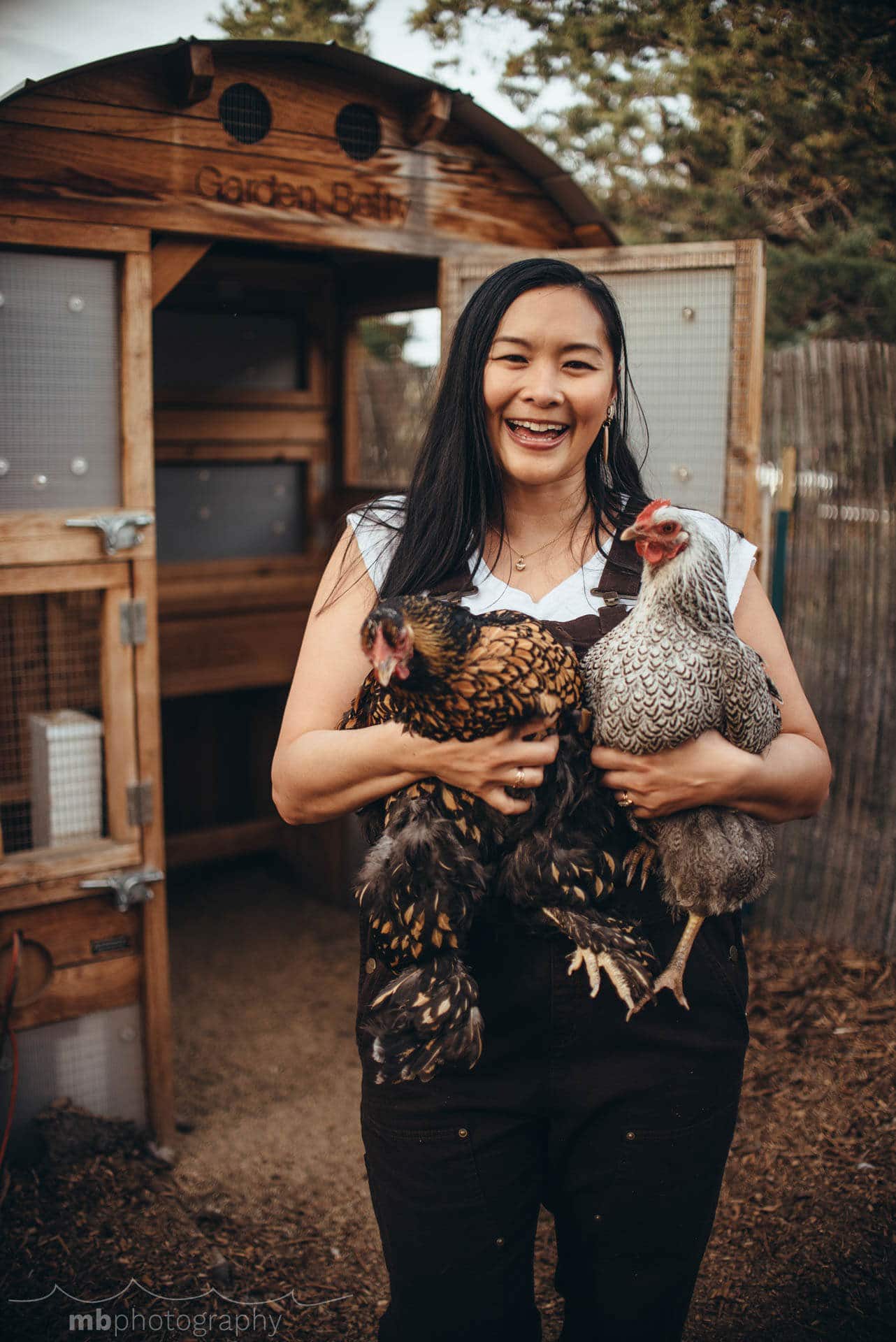 Garden Betty holding two chickens in front of their coop
