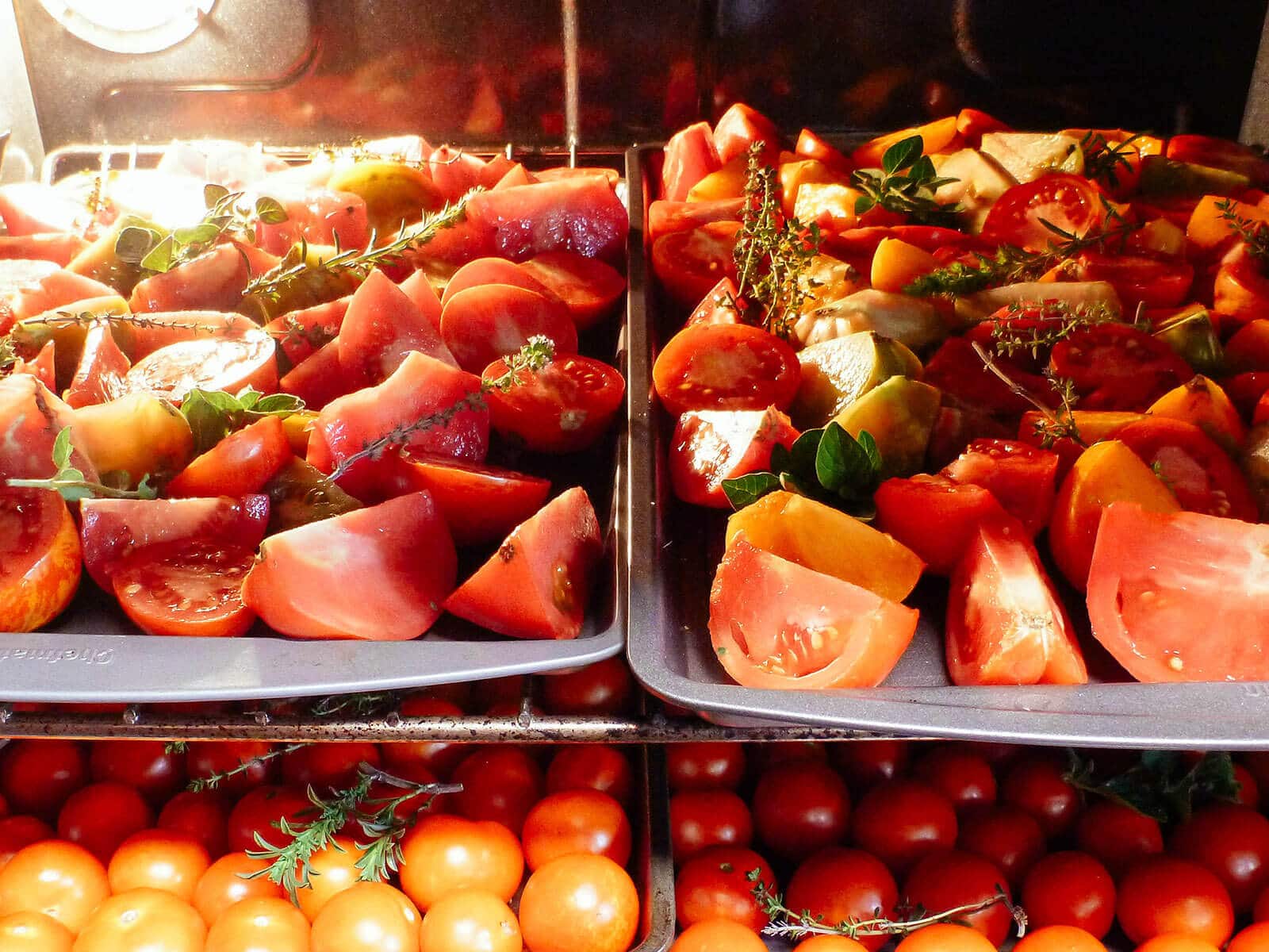 Four baking sheets filled with whole cherry tomatoes, tomato slices, and tomato wedges being dried in the oven