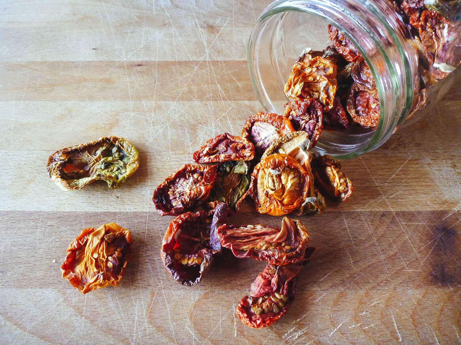 Colorful oven-dried tomatoes spilling out of a glass jar on a butcher block