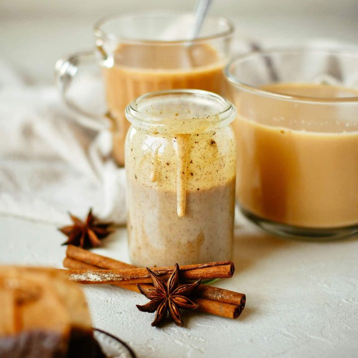 A jar of homemade chai concentrate sitting in front of two mugs of chai, with cinnamon sticks, star anise, and brewed tea bags in the foreground